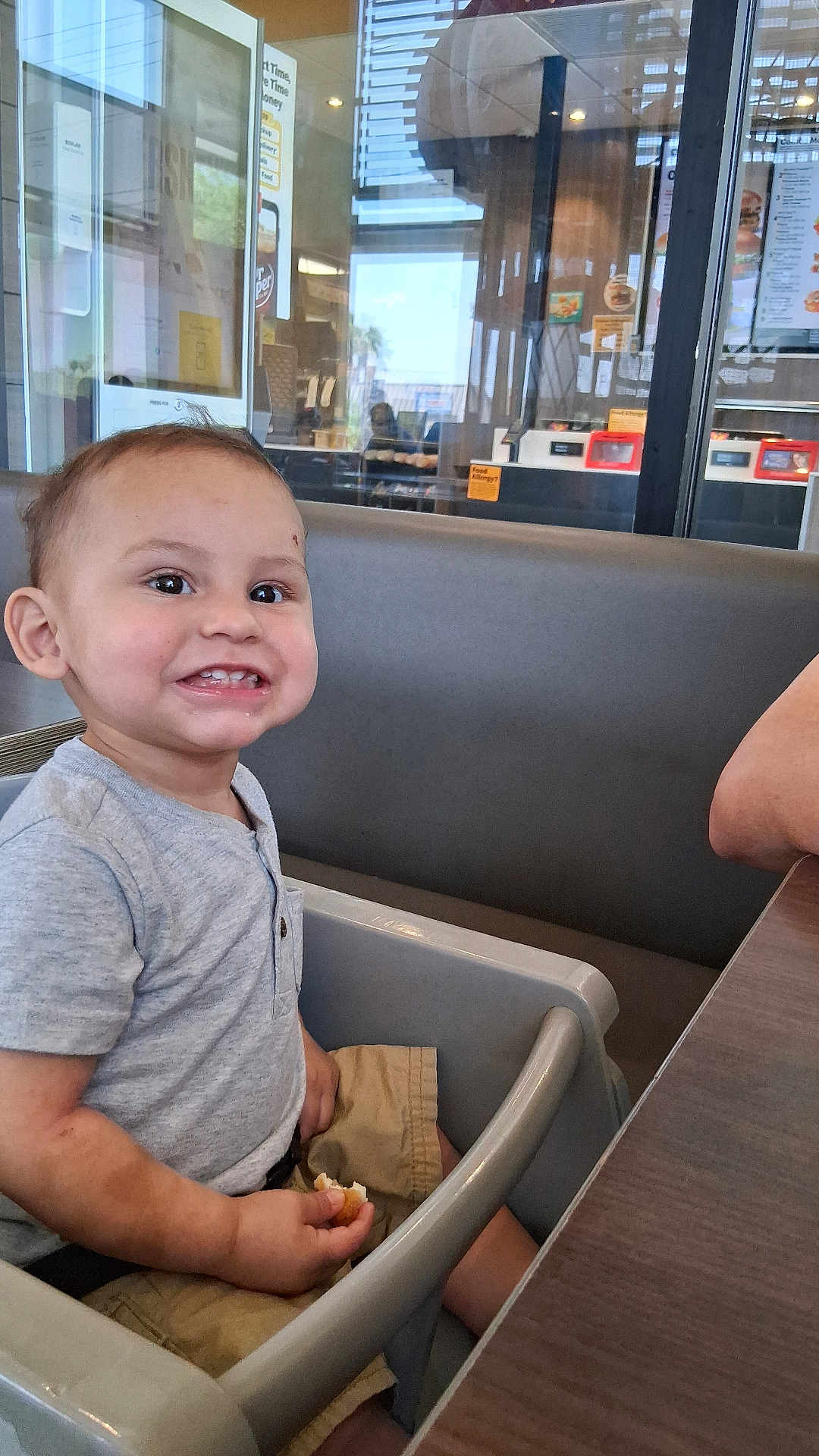 Noah is registered to the contest to win money with this photo: toddler, child, high_chair, smiling, food, indoor, restaurant, table, seat, casual, menu, reflection, window, shorts, shirt, person, happy, baby, cute, dining