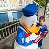 child, donald_duck, costume, autograph, superhero_outfit, outdoor, porch, flowers, greenery, sidewalk, character, theme_park, childhood, happy, person, signature, blue_hat, white_gloves, cartoon_character, daytime