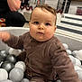 baby, child, ball_pit, plastic_balls, brown_clothing, indoor, festive_decorations, curious_expression, playful, person, face, head, seated, toy, blurred_motion, stairs, railing, adult_arm, background, home