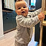 Izaac participe au concours pour gagner de l'argent avec cette photo : toddler, child, baby, standing, indoors, kitchen, wooden_furniture, gray_clothing, socks, floor, window, cabinet, curious, face, hair, mocha_hair, expression, person, home, light