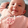 baby, smile, pink_sweater, pacifier, chain, lying_down, cozy, blanket, face, tongue, cute, infant, child, soft_texture, warmth, indoors, happy, closeup, head, person