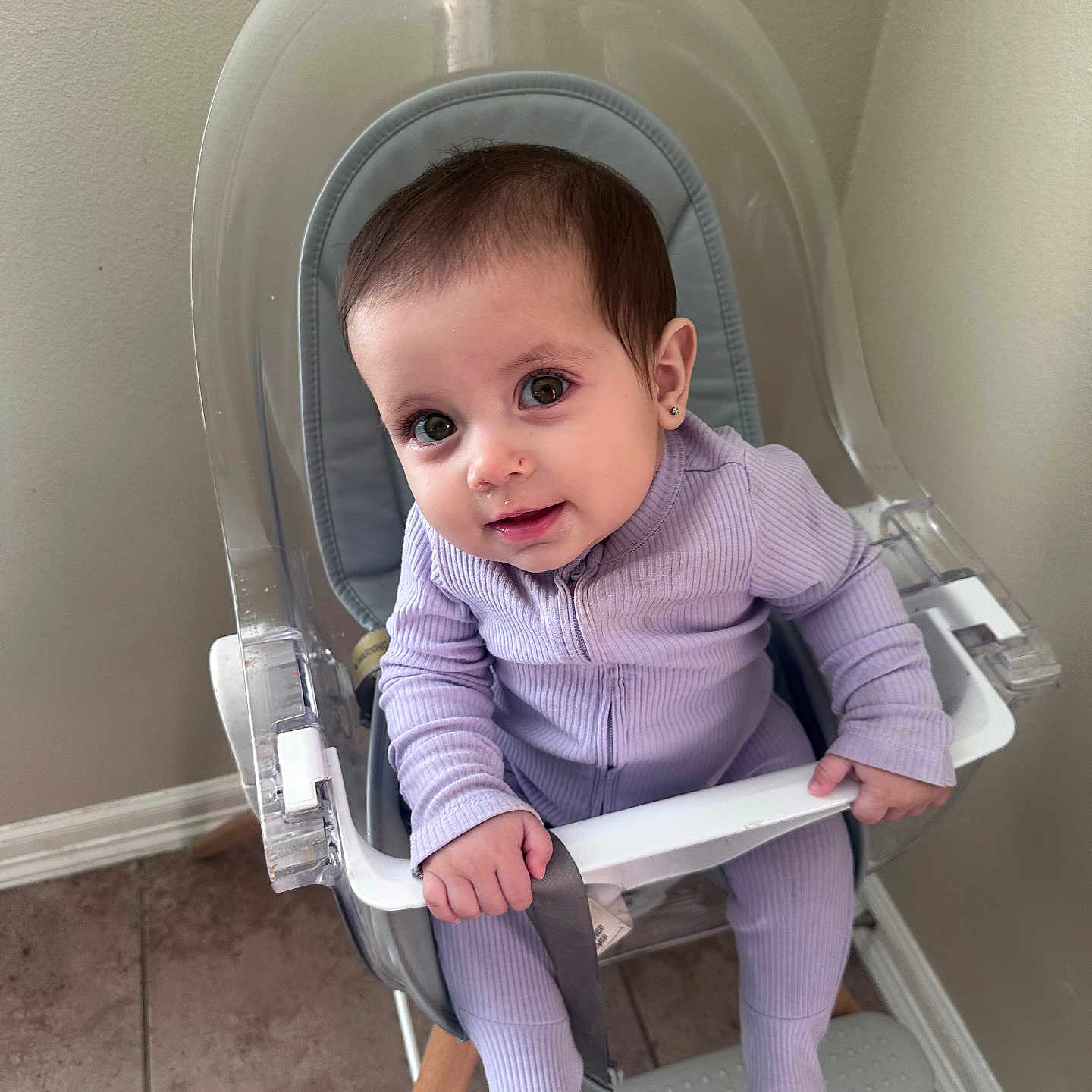 Isabella joined the competition — help win amazing prizes! baby, child, curious, earrings, floor, footrest, grip, high_chair, home, indoor, modern_furniture, person, purple_onesie, safety_bar, sitting, smiling, tile_floor, transparent_chair, wall, young_child