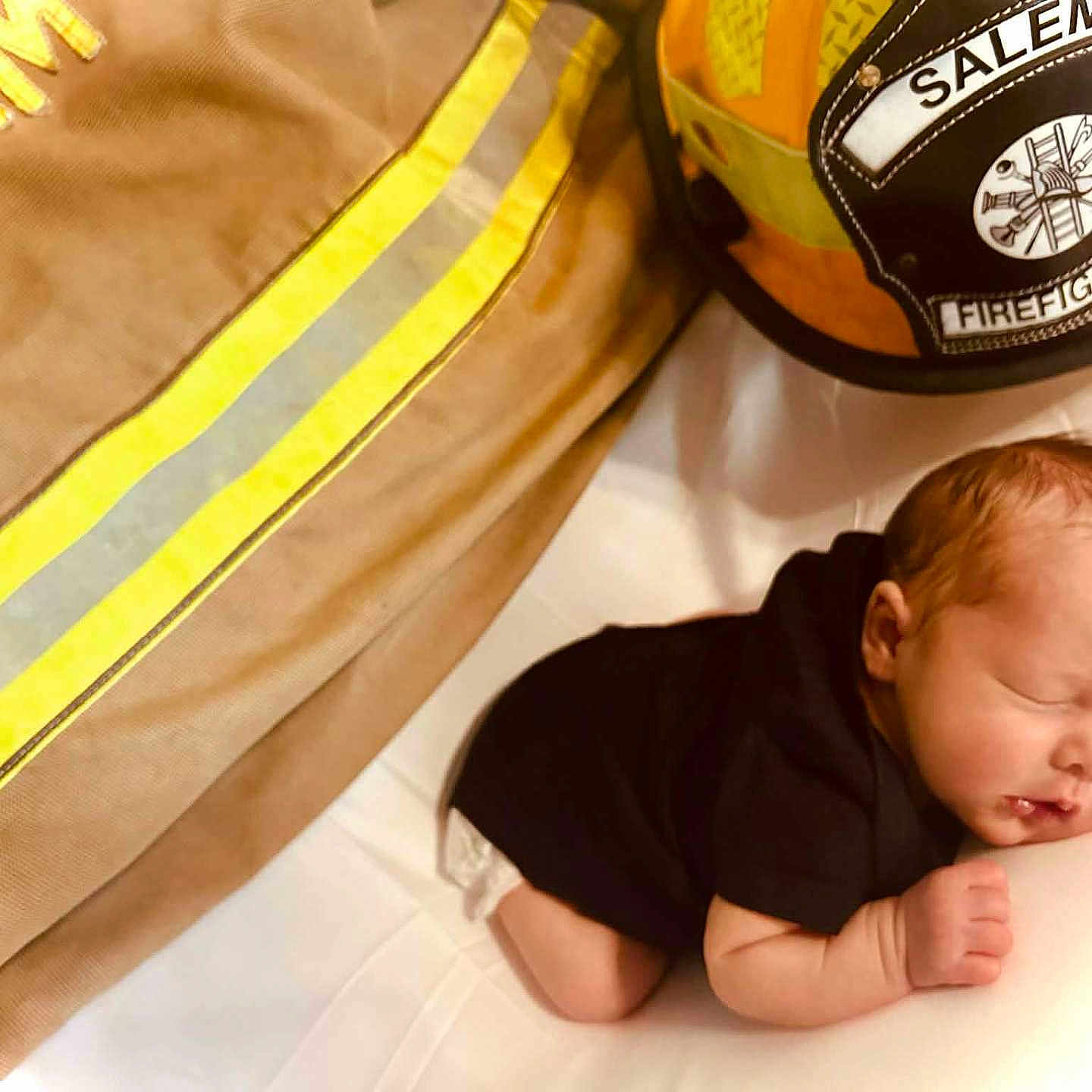 Keegan is registered to the contest to win money with this photo: baby, newborn, sleeping, firefighter, helmet, uniform, black_clothing, peaceful, infant, cute, resting, portrait, closeup, human, skin, head, hand, bed, white_background, soft