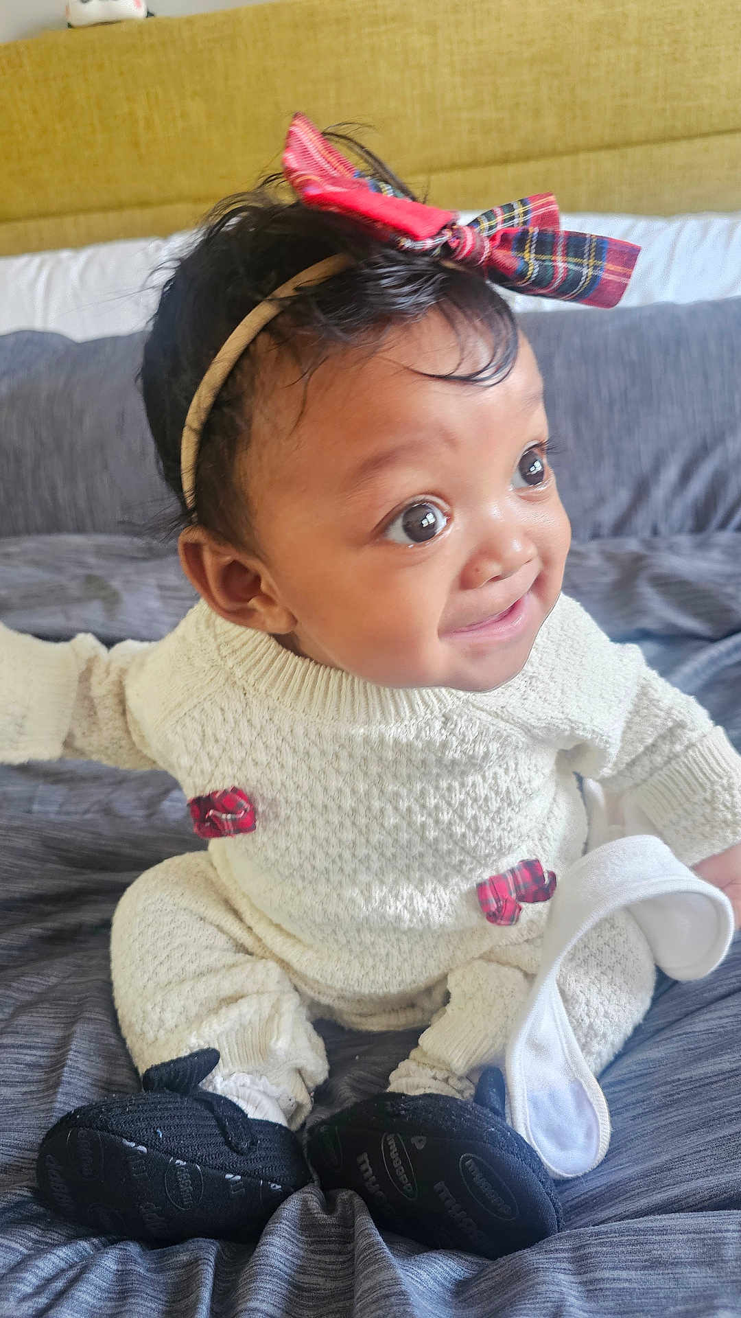 Caleigh is registered to the contest to win money with this photo: baby, child, infant, knitwear, headband, bow, plaid, sock, footwear, bed, blanket, smiling, sitting, indoors, cute, young_child, black_hair, cozy, apparel, portrait