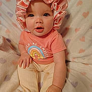 Maci joined the competition — help win amazing prizes! baby, child, clothing, cute, expression, face, footed_pants, hand, happy, heart_pattern, indoors, infant, pastel_colors, pink_bonnet, portrait, rainbow_shirt, sitting, smiling, soft_blanket, young_child