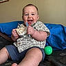 baby, child, smiling, happy, blue_eyes, snack, shirt, shorts, toy, green_ball, bed, blanket, foot, hand, indoor, wall, picture_frame, person, infant, casual_clothing