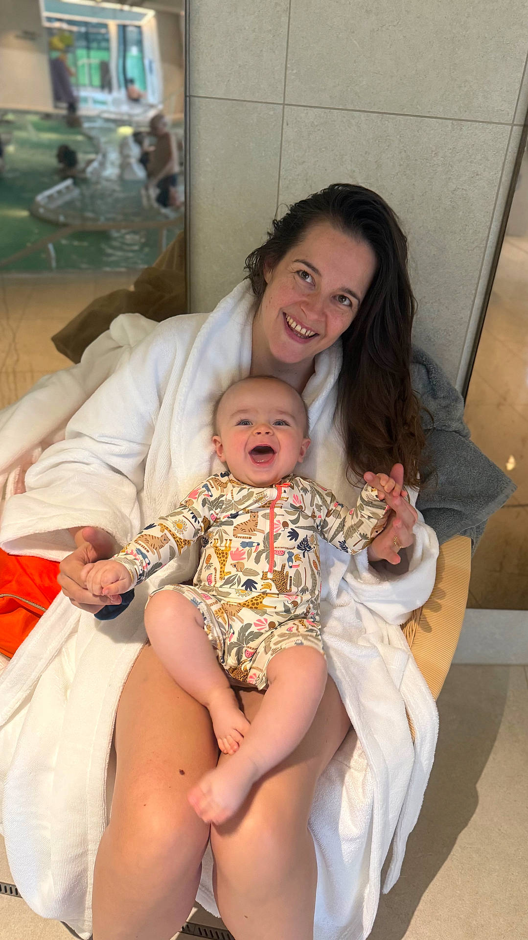 Isaïa a rejoint le concours — aidez-le/la à gagner de superbes lots ! animal_print, baby, bathrobe, blurred_background, casual, child, face, happy, holding_hands, indoor, joyful, legs, long_hair, person, pool_area, relaxed, seated, skin, smiling, woman