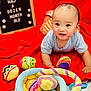 baby, infant, child, toys, plush, red_blanket, colorful, smiling, face, blue_shirt, orange_shorts, wooden_board, text_board, indoor, cute, playtime, happy, soft_toys, fabric, portrait