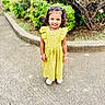 child, girl, yellow_dress, smile, outdoor, pavement, greenery, bushes, bracelets, necklace, hair_clips, curly_hair, happy, standing, footwear, sunny, daytime, nature, portrait, casual