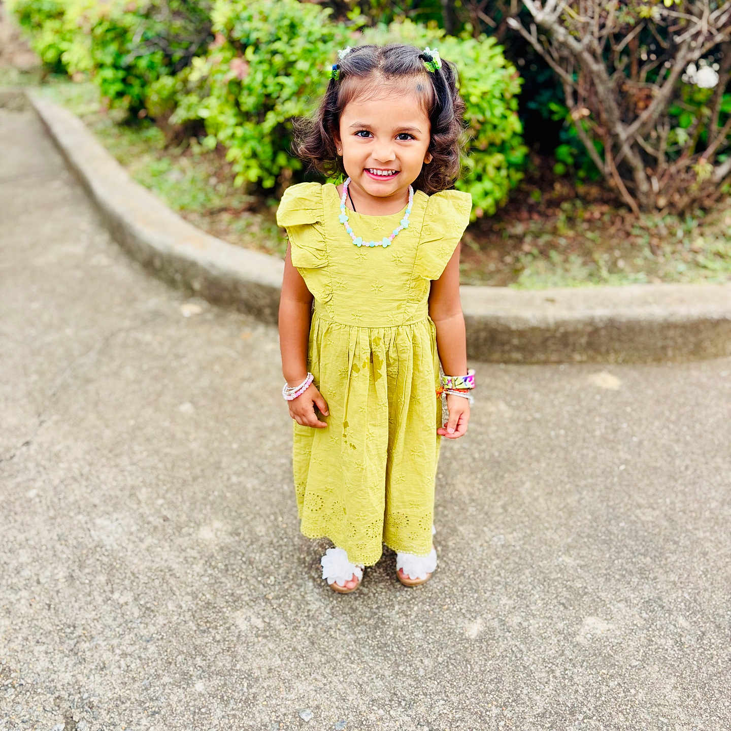 Veda joined the competition — help win amazing prizes! child, girl, yellow_dress, smile, outdoor, pavement, greenery, bushes, bracelets, necklace, hair_clips, curly_hair, happy, standing, footwear, sunny, daytime, nature, portrait, casual