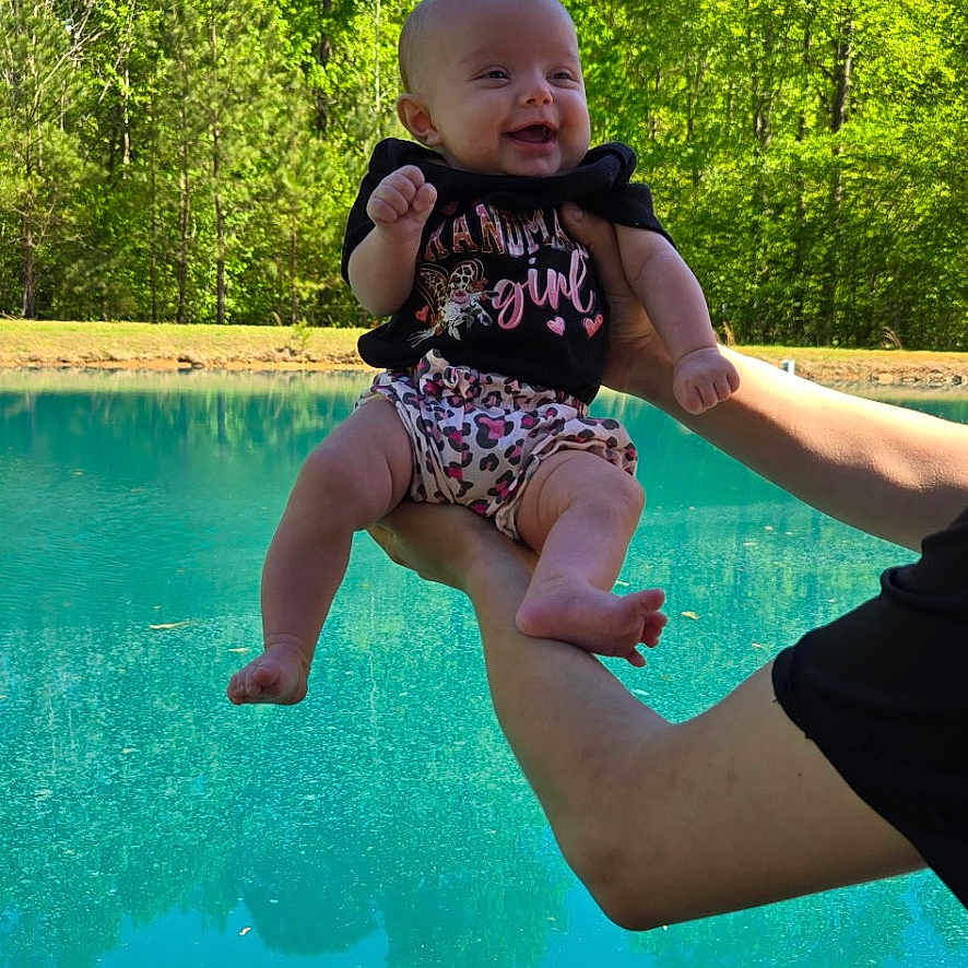 Sadie joined the competition — help win amazing prizes! arms, baby, child, clothing, cute, greenery, happy, holding, infant, legs, nature, outdoor, person, pond, shorts, smiling, sunlight, trees, tshirt, water
