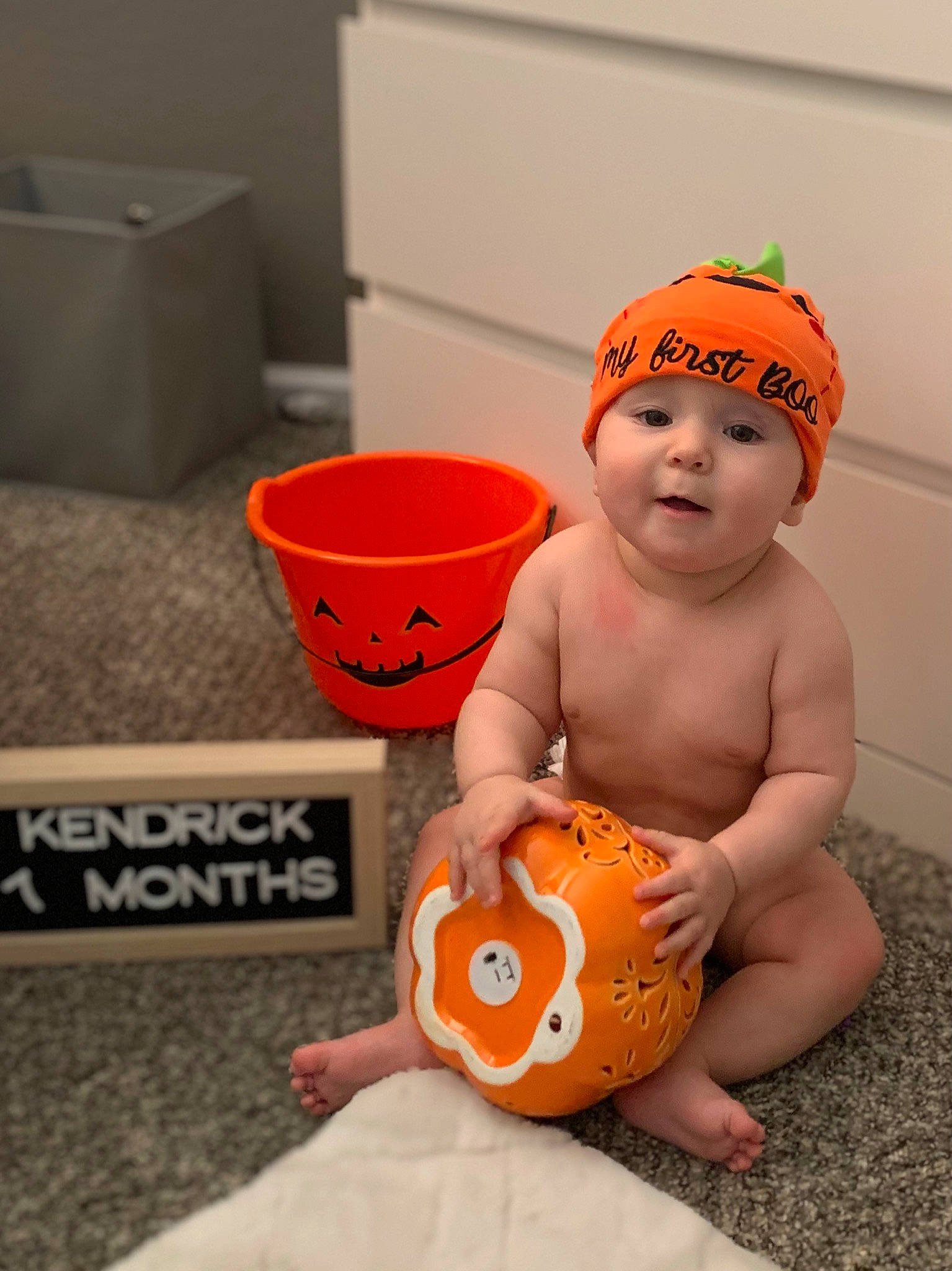 Kendrick is registered to the contest to win money with this photo: baby, bucket, calabaza, cap, child, floor, flooring, fun, goggles, happy, headwear, human_body, musical_instrument, orange, person, personal_protective_equipment, play, room, sitting, stomach
