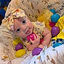 baby, infant, child, headband, tongue_out, blue_eyes, plastic_eggs, toy, blanket, patterned, cute, playful, hand, clothing, yellow, purple, pink, blue, soft_toys, smiling