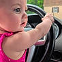 baby, child, infant, face, arm, hand, steering_wheel, car_interior, pink_clothing, polka_dot, closeup, portrait, curious, cute, vehicle, dashboard, window, green_background, seat, person