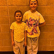 Kiera is registered to the contest to win money with this photo: children, face_paint, brick_wall, indoor, socks, sweatpants, tshirt, smiling, standing, floor, headband, casual_clothing, two_people, portrait, child, kids, happy, wall, footwear, young