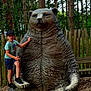 child, boy, statue, bear, forest, trees, wooded_area, outdoor, nature, summer, cap, shorts, tshirt, fence, bench, playground, park, footwear, expression, daytime