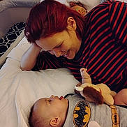 Christopher James joined the competition — help win amazing prizes! baby, woman, red_hair, batman_outfit, toy, bed, blanket, smile, face, infant, clothing, indoor, person, head, cuddly_toy, happy, interaction, child, parent, playful