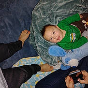 Christopher James joined the competition — help win amazing prizes! baby, blanket, blue, carpet, casual, child, clothing, comfort, cushion, feet, green_shirt, hand, happy, indoor, lying_down, person, phone, smile, stuffed_animal, toy