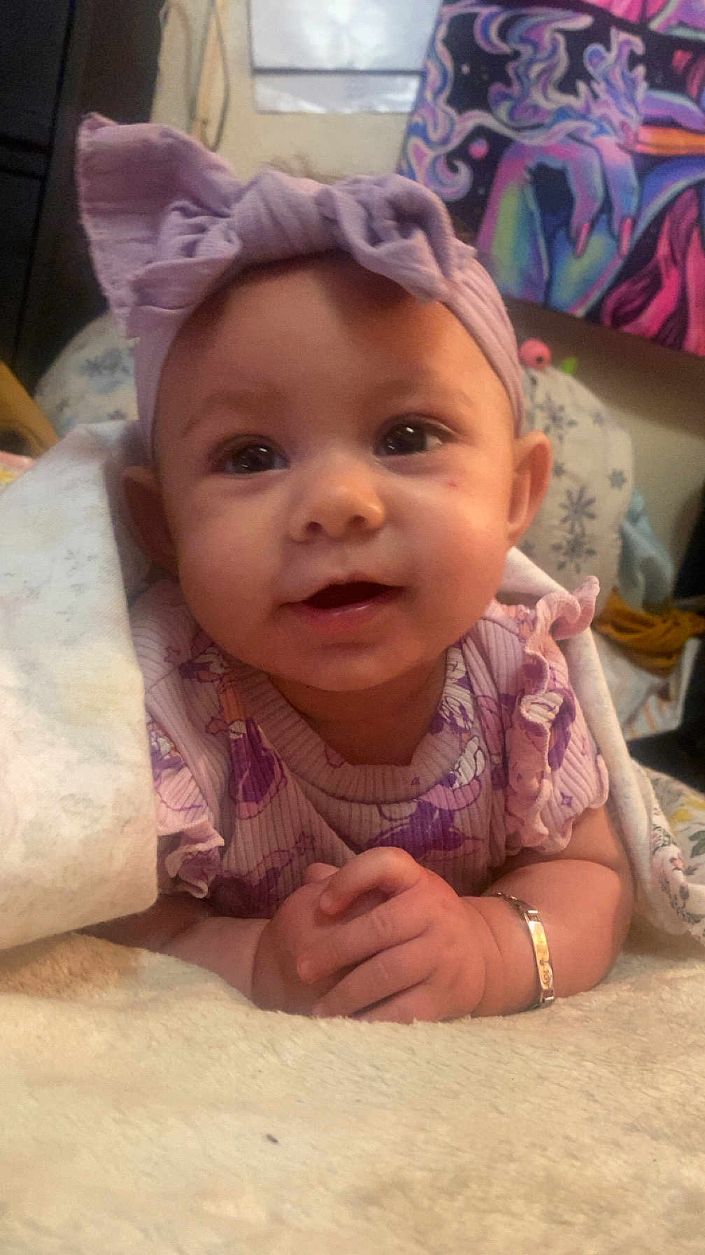 Sedona is registered to the contest to win money with this photo: baby, infant, headband, purple_clothing, blanket, smiling, face, hands, bracelet, indoors, cute, child, portrait, soft_texture, cozy, patterned_background, happy, lying_down, young_child, home