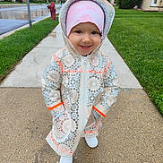Mia is registered to the contest to win money with this photo: child, toddler, smile, raincoat, hood, hat, sidewalk, wet_sidewalk, grass, neighborhood, row_houses, fire_hydrant, rain, shoes, portrait, outdoors, cute, happy, pavement, street
