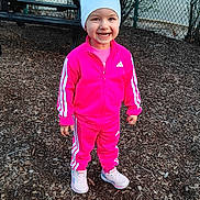 Mia joined the competition — help win amazing prizes! athletic_wear, casual_clothing, child, cute, daylight, fence, happy, nature, outdoor, person, picnic_table, pink_tracksuit, playground, smiling, sneakers, sportswear, standing, white_cap, wood_chips, young_child