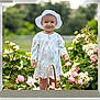 child, baby, toddler, smiling, sun_hat, dress, floral_dress, basket, pink_flowers, white_flowers, garden, outdoors, greenery, portrait, standing, cute, happy, petals, foliage, lawn