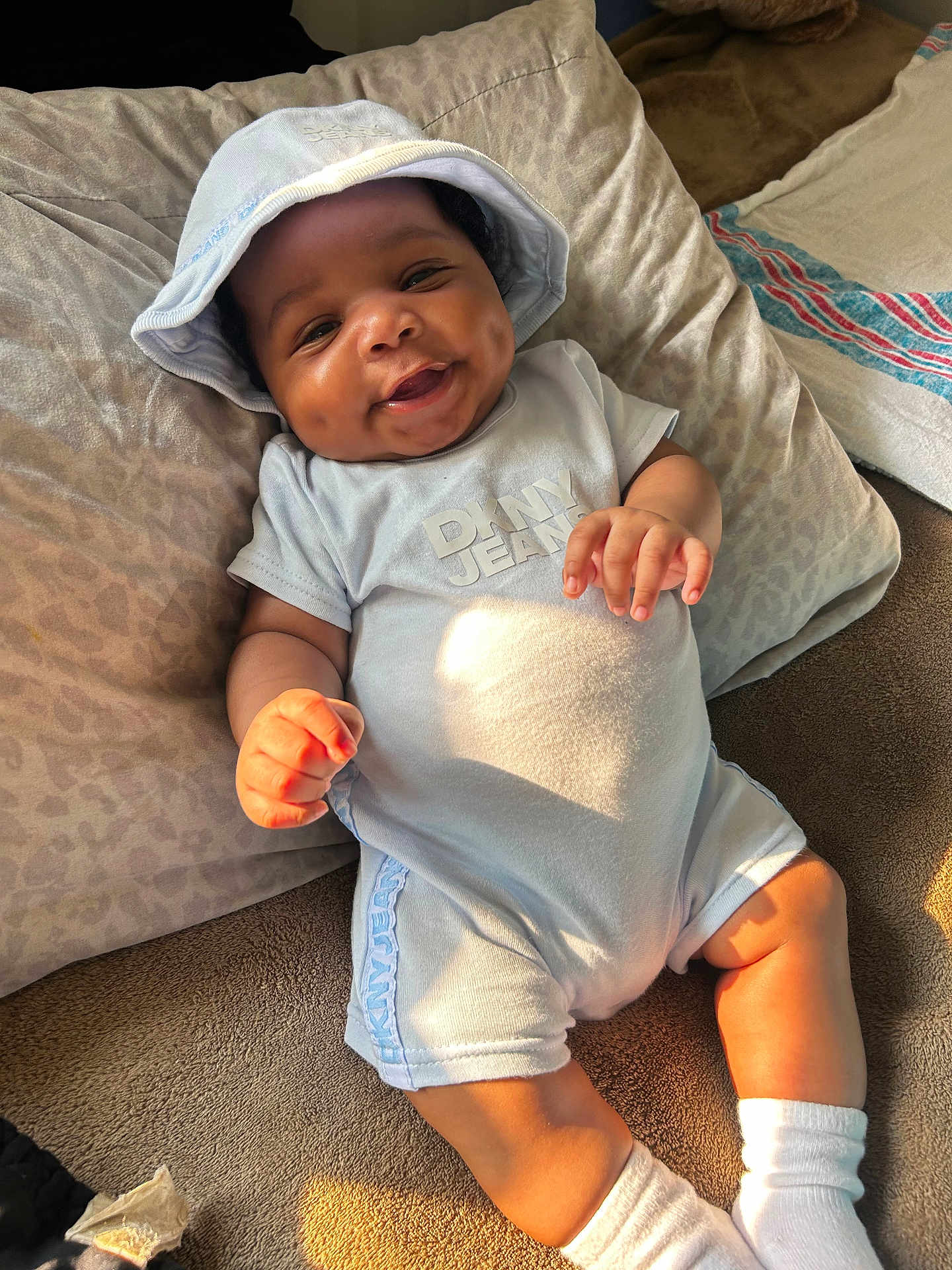 Deomari is registered to the contest to win money with this photo: baby, smiling, dimples, hat, clothing, light_blue, socks, bed, pillow, sunlight, infant, cute, happy, indoor, portrait, child, toddler, comfortable, soft, relaxed