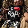 baby, child, smiling, dimples, curly_hair, tshirt, shorts, camouflage, text_on_clothing, pillow, blanket, indoor, cozy, relaxing, portrait, cute, person, infant, happy, seated