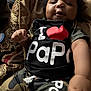 baby, child, infant, camouflage_clothing, shirt, shorts, cushion, patterned_fabric, indoor, person, face, hand, text_on_clothing, love_heart, relaxing, seated, cute, soft_lighting, closeup, portrait