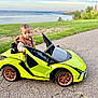 toddler, child, toy_car, green_car, outdoor, grass, water, road, path, asphalt, nature, vehicle, park, casual_clothing, vest, expression, summer, daytime, wheels, small_car