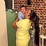 baby, woman, smiling, kissing, yellow_dress, striped_clothes, long_hair, brick_wall, flowers, indoor, love, happy, holding, portrait, family, child, person, fashion, casual, background
