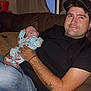 Jesse is registered to the contest to win money with this photo: baby, bracelet, cap, couch, curtain, face, hand, holding, home, indoor, infant, jeans, man, pajamas, person, relaxing, seated, smile, tattoo, window