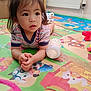 Angela is registered to the contest to win money with this photo: toddler, child, baby_bottle, play_mat, indoor, floor, radiator, colorful, striped_clothing, curly_hair, toy, cartoon, animal_print, food_print, hand, face, expression, person, home, crawling