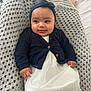 baby, infant, headband_bow, navy_cardigan, white_dress, knitted_blanket, crochet_blanket, bed, smile, face, chubby_cheeks, hands, eyes, portrait, indoors, cozy, cute, lying_down, fabric_texture, soft_light