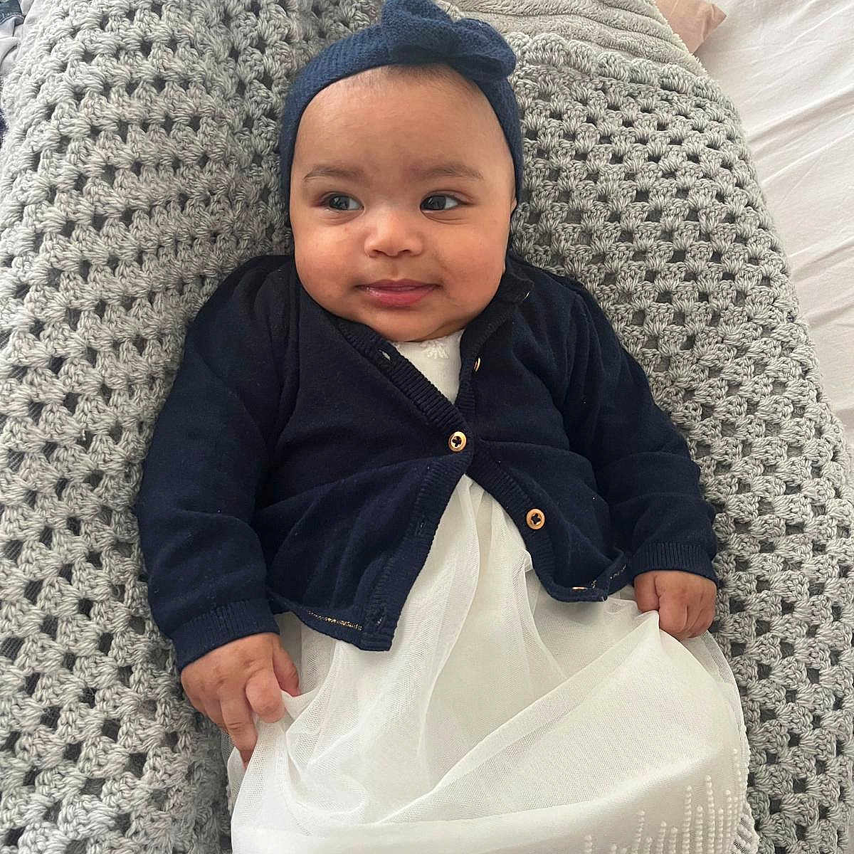 Elyssia a rejoint le concours — aidez-le/la à gagner de superbes lots ! baby, bed, chubby_cheeks, cozy, crochet_blanket, cute, eyes, fabric_texture, face, hands, headband_bow, indoors, infant, knitted_blanket, lying_down, navy_cardigan, portrait, smile, soft_light, white_dress