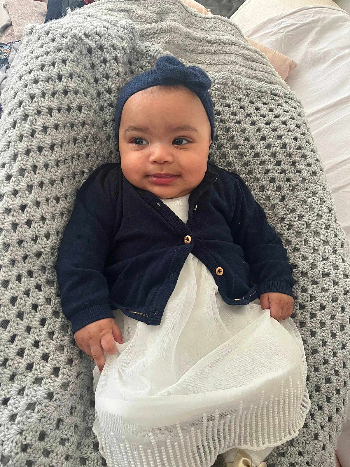 Elyssia a rejoint le concours — aidez-le/la à gagner de superbes lots ! baby, infant, headband_bow, navy_cardigan, white_dress, knitted_blanket, crochet_blanket, bed, smile, face, chubby_cheeks, hands, eyes, portrait, indoors, cozy, cute, lying_down, fabric_texture, soft_light