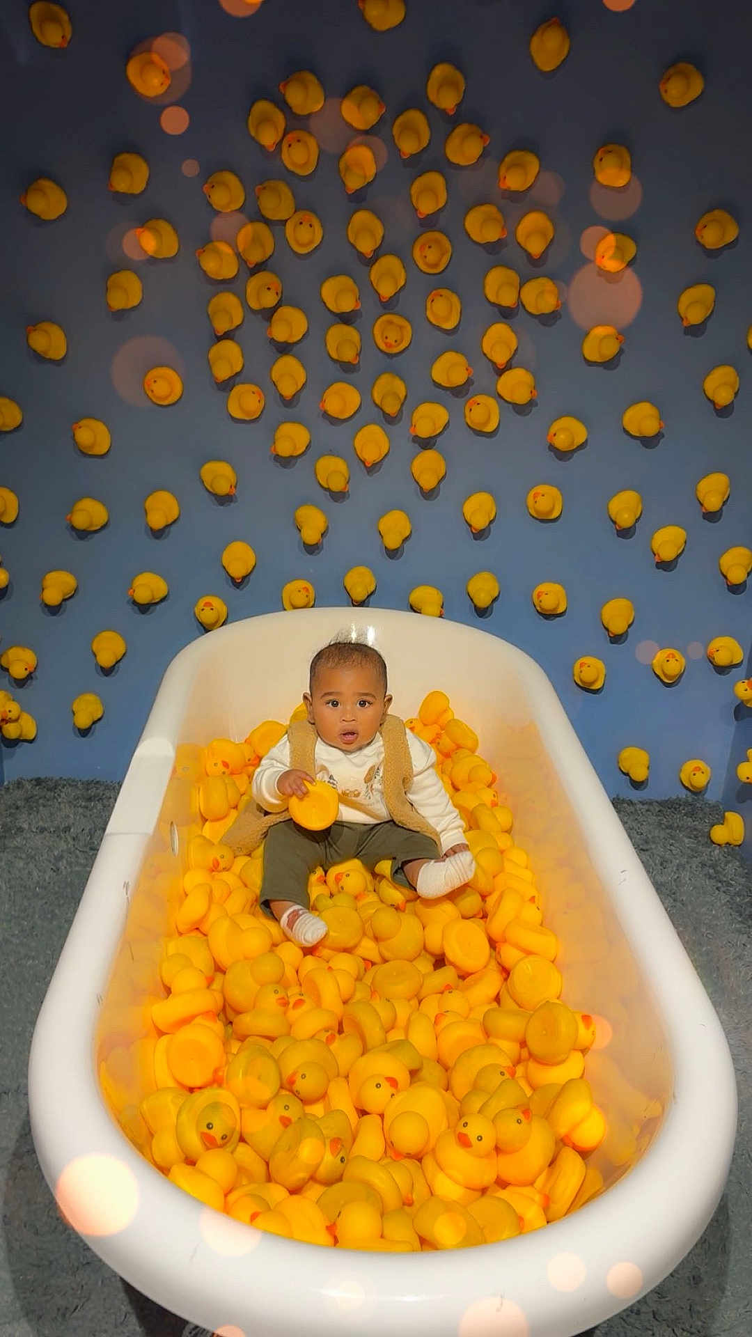 Zane joined the competition — help win amazing prizes! baby, bathtub, rubber_duck, yellow, toy, child, indoor, curious, sitting, blue_wall, playful, cute, soft_lighting, floor_rug, clothing, vest, white_socks, expression, portrait, decor