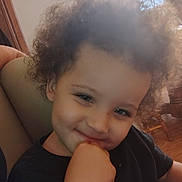 Jayden is registered to the contest to win money with this photo: child, toddler, curly_hair, smiling, portrait, close_up, indoor, home, couch, window, blinds, wooden_floor, black_shirt, face, eyes, nose, mouth, hand, seated, candid
