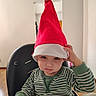 child, toddler, santa_hat, christmas, portrait, sitting, high_chair, striped_shirt, green_clothing, white_trim, blue_eyes, serious_expression, indoor, home, chair, table, baby, festive, holiday, photography