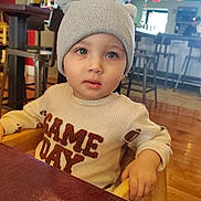 Jayden is registered to the contest to win money with this photo: baby, child, infant, portrait, sitting, restaurant, table, beanie, hat, pom_pom, sweater, clothing, blue_eyes, wide_eyed, looking_at_camera, indoor, bar, neon_sign, wooden_floor, smiling