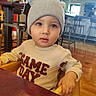 baby, child, infant, portrait, sitting, restaurant, table, beanie, hat, pom_pom, sweater, clothing, blue_eyes, wide_eyed, looking_at_camera, indoor, bar, neon_sign, wooden_floor, smiling