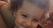 Jayden is registered to the contest to win money with this photo: child, toddler, curly_hair, smiling, portrait, close_up, face, eyes, indoor, home, living_room, couch, window, blinds, wooden_floor, black_shirt, seated, hand_on_chin, warm_lighting, photograph