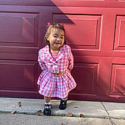 Kiana is registered to the contest to win money with this photo: child, girl, pink_clothing, checkered_pattern, smiling, hair_accessories, shoes, sidewalk, autumn_leaves, outdoor, garage_door, concrete, fall, cute, portrait, standing, casual, daylight, happy, young