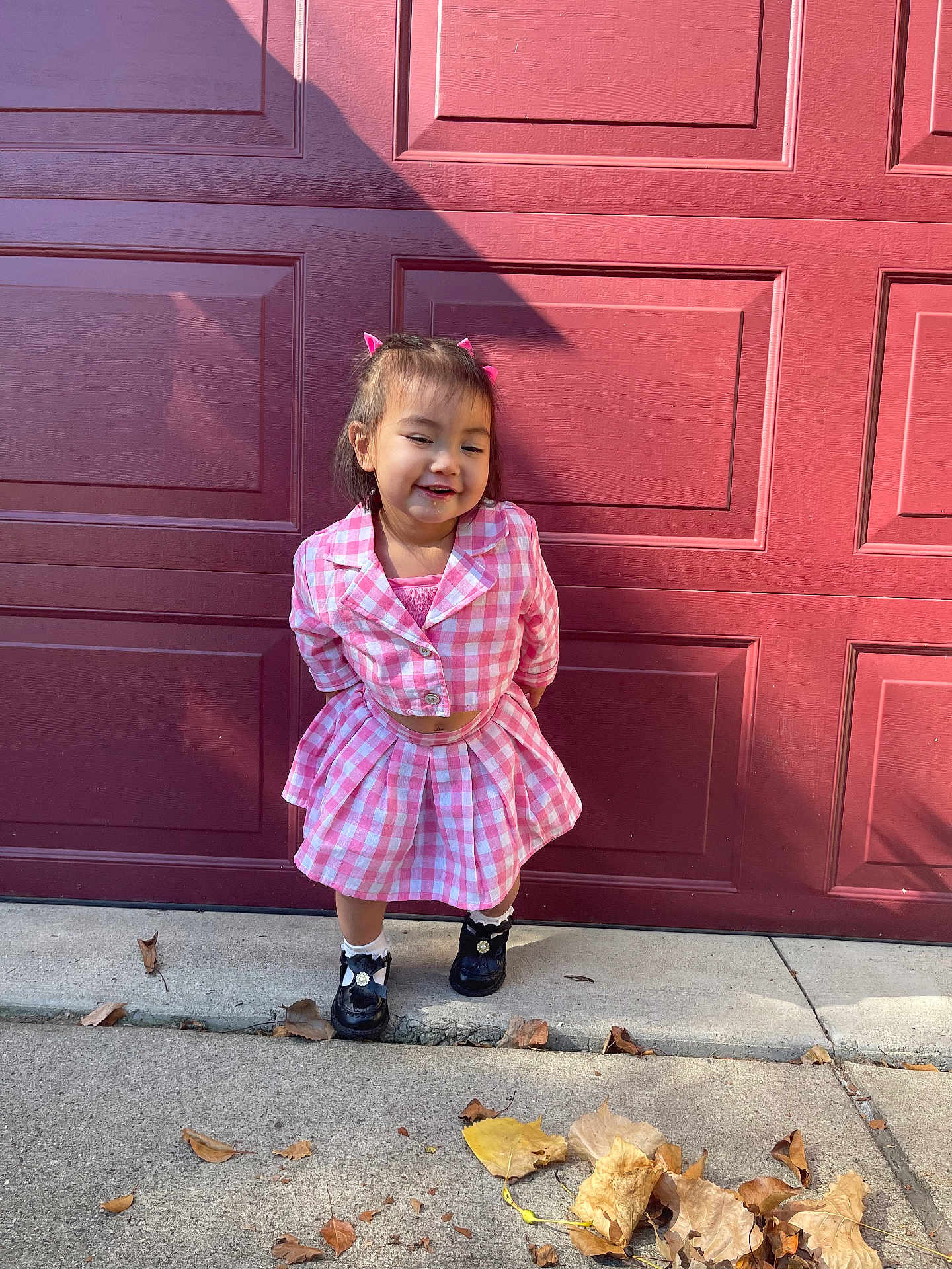 Kiana is registered to the contest to win money with this photo: child, girl, pink_clothing, checkered_pattern, smiling, hair_accessories, shoes, sidewalk, autumn_leaves, outdoor, garage_door, concrete, fall, cute, portrait, standing, casual, daylight, happy, young
