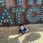Lucca is registered to the contest to win money with this photo: child, toddler, straw, crawling, wood, letters, lighting, shadow, outdoor, sign, curious, young, person, casual_clothing, daylight, background, playful, illuminated_letters, brown, blue_shirt