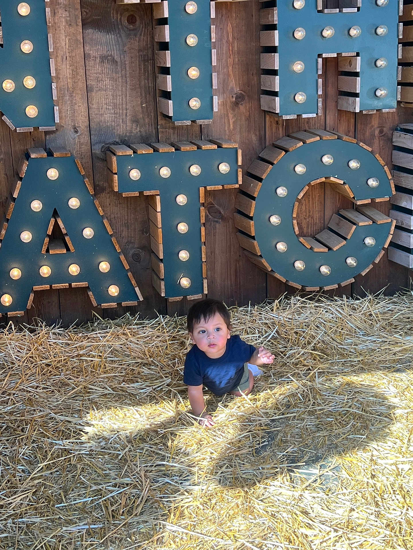 Lucca is registered to the contest to win money with this photo: child, toddler, straw, crawling, wood, letters, lighting, shadow, outdoor, sign, curious, young, person, casual_clothing, daylight, background, playful, illuminated_letters, brown, blue_shirt