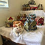 cat, orange_cat, white_cat, miniature_houses, christmas_village, table, white_cloth, window, indoor, decorations, holiday, cozy, feline, pet, resting, home_decor, festive, ornaments, vintage, scene