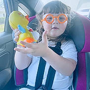Ruby joined the competition — help win amazing prizes! child, toddler, car_seat, toy, duck, glasses, orange_glasses, seatbelt, car_interior, window, suburban, daylight, curly_hair, white_shirt, purple_car_seat, plastic_toy, happy, indoor, person, smiling