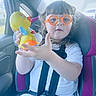 car_interior, car_seat, child, curly_hair, daylight, duck, glasses, happy, indoor, orange_glasses, person, plastic_toy, purple_car_seat, seatbelt, smiling, suburban, toddler, toy, white_shirt, window