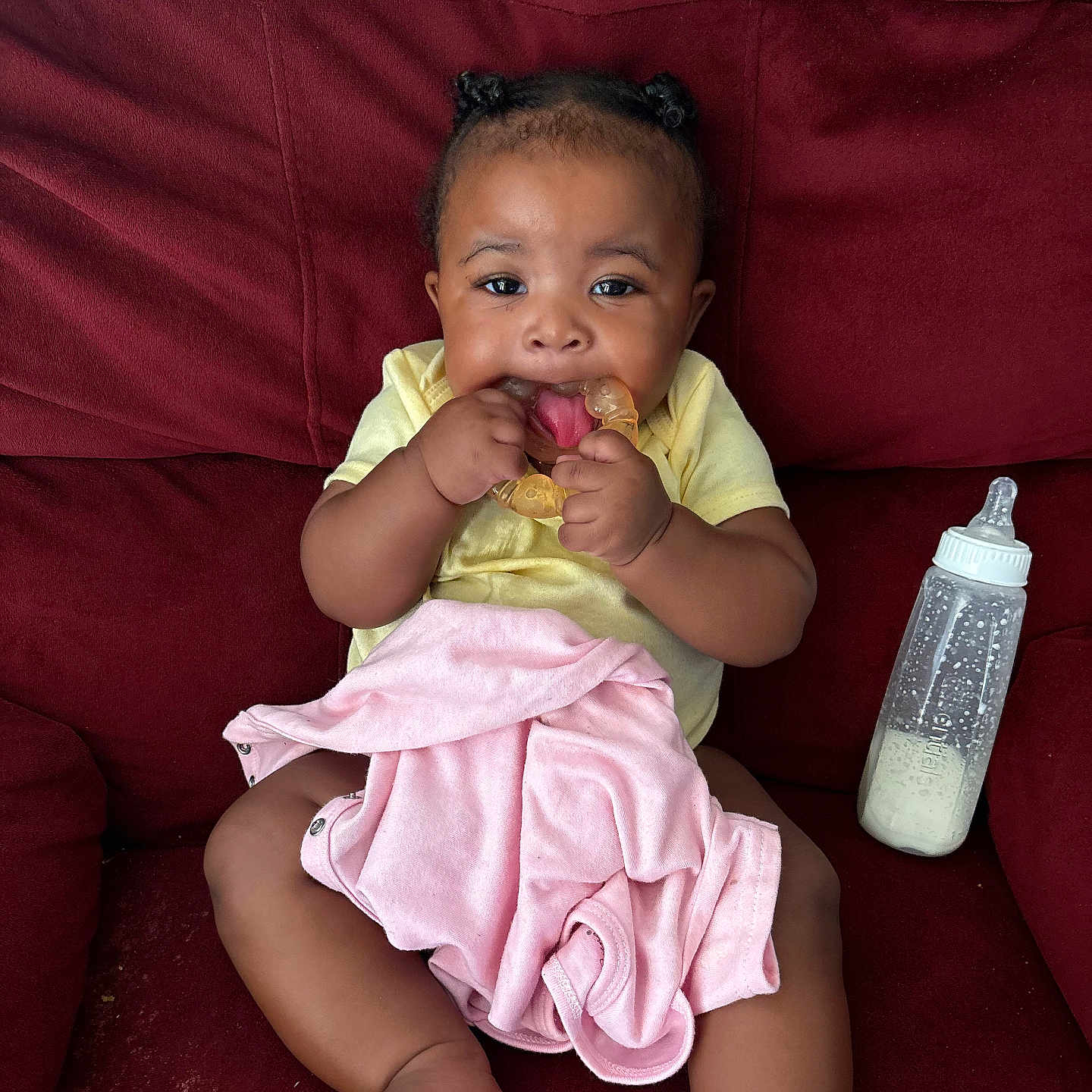 Niari joined the competition — help win amazing prizes! baby, infant, teething_ring, blanket, bottle, milk, couch, red_couch, clothing, yellow_shirt, cute, child, person, indoors, sitting, happy, playful, small_hands, pigtails, portrait